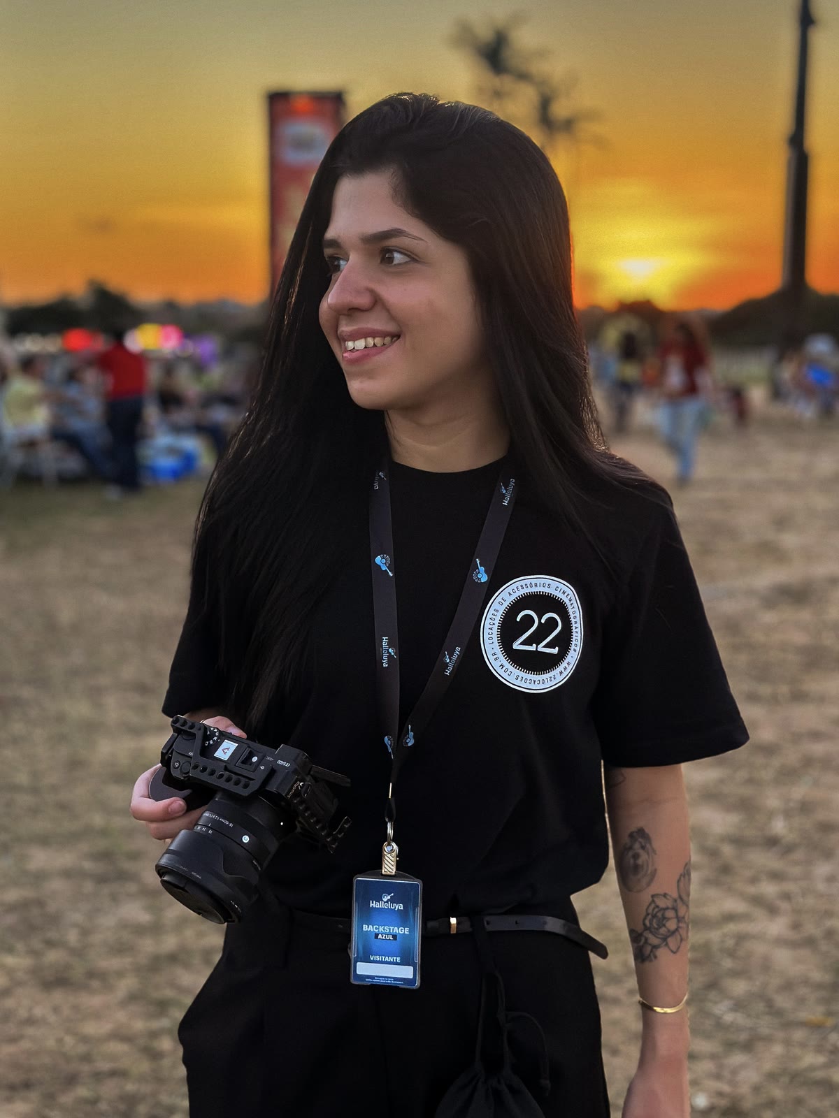 Sarah Aragão em um evento ao entardecer, câmera na mão e crachá de bastidores no pescoço.