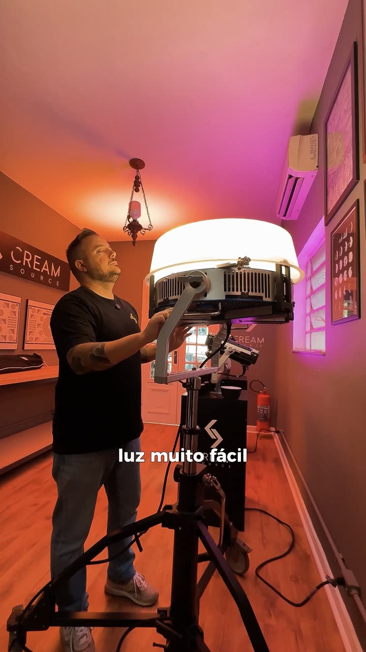Operador ajustando um painel de LED grande em tripé, em sala com luz rosa e azul.